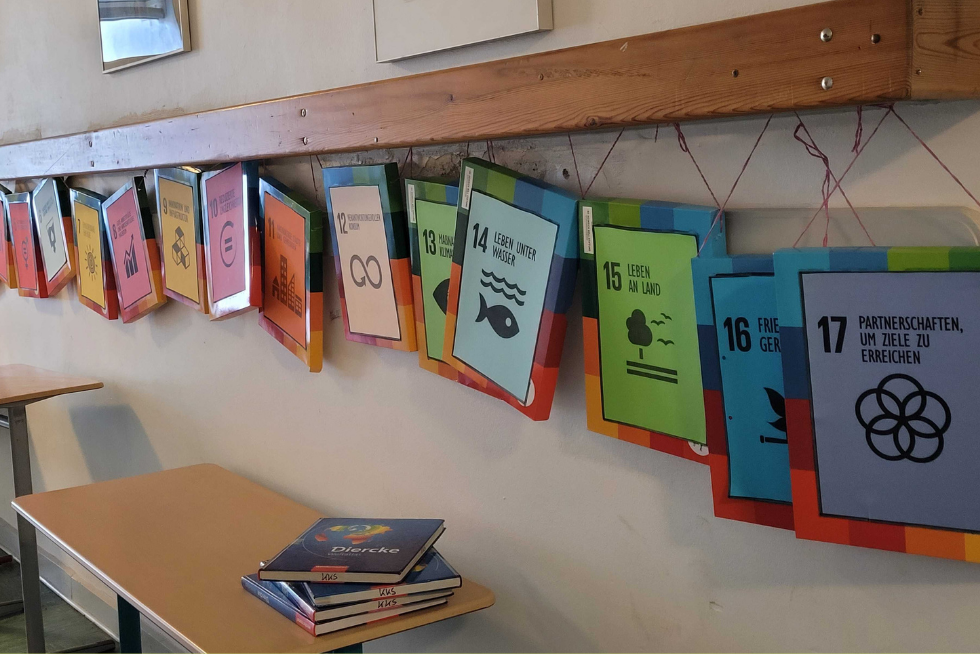 SDGs der UN An einer langen Garderobe aus Holz hängen bunte Schilder. Auf den Schildern stehen jeweils die Zahlen von 1 bis 17 und jeweils ein Nachhaltigkeitsziel der UN.