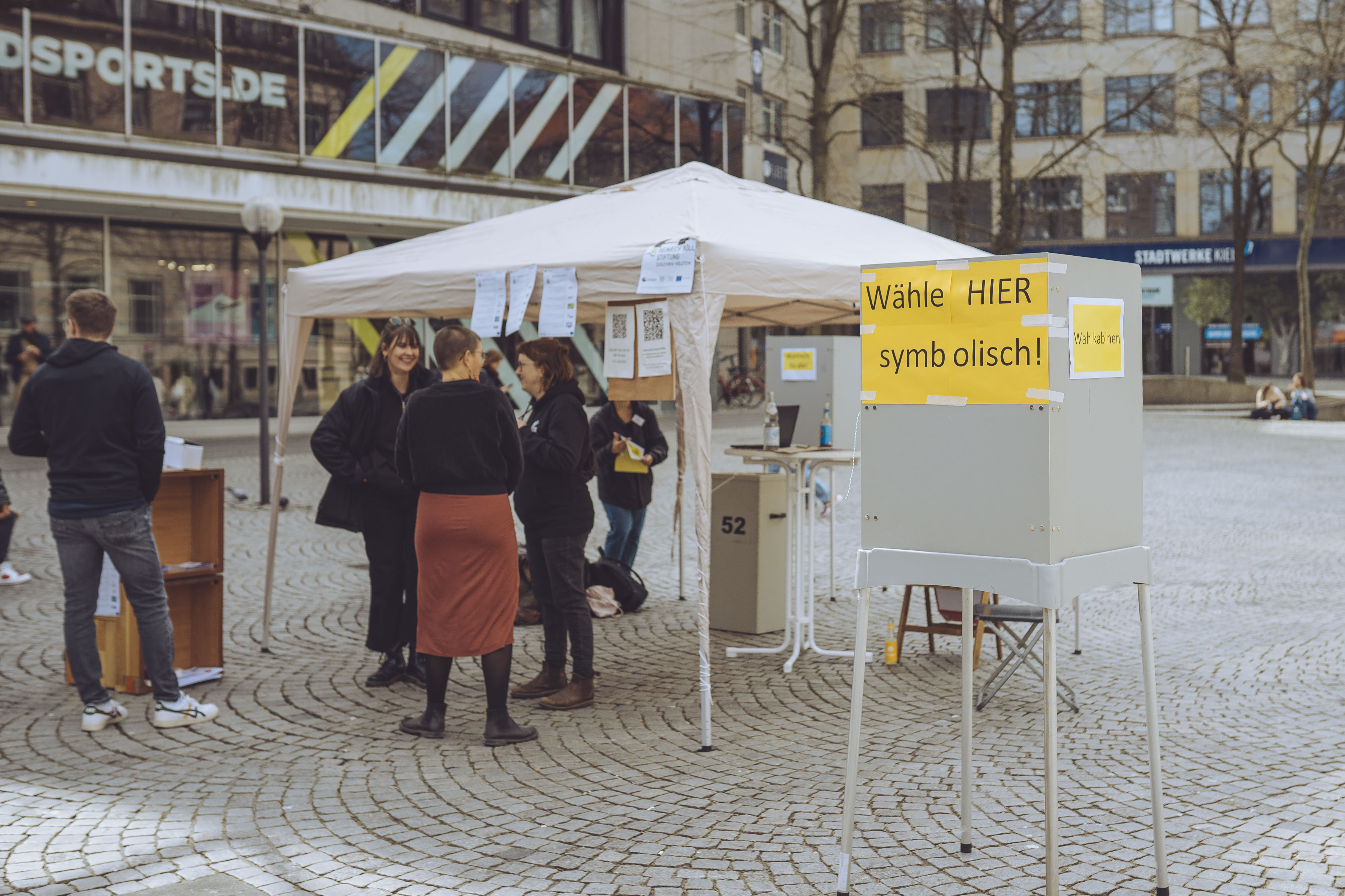 Symbolische Wahlen 2022 Auf einem Platz steht vorne eine Wahlkabine, auf der steht: Wähle hier symbolisch. Dahinter ist ein Pavillon aufgebaut, vor dem eine Gruppe von Menschen steht.