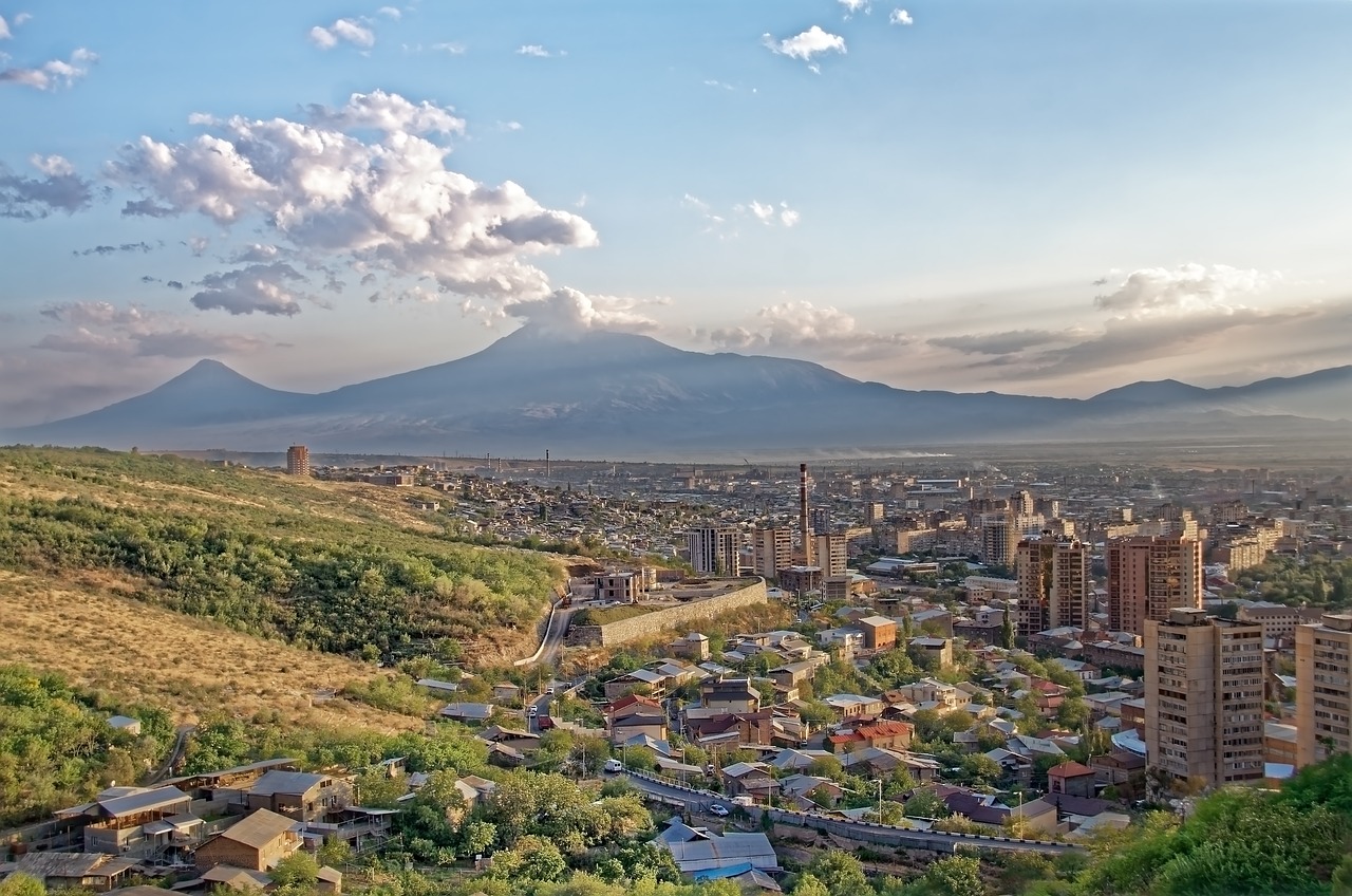 Die armenische Hauptstadt Yerevan aus der Vogelperspektive. Sie liegt eingebettet in Hügel, einige Hochäuser ragen aus der Stadt heraus. Am Horizont sind Berge und Wolken.