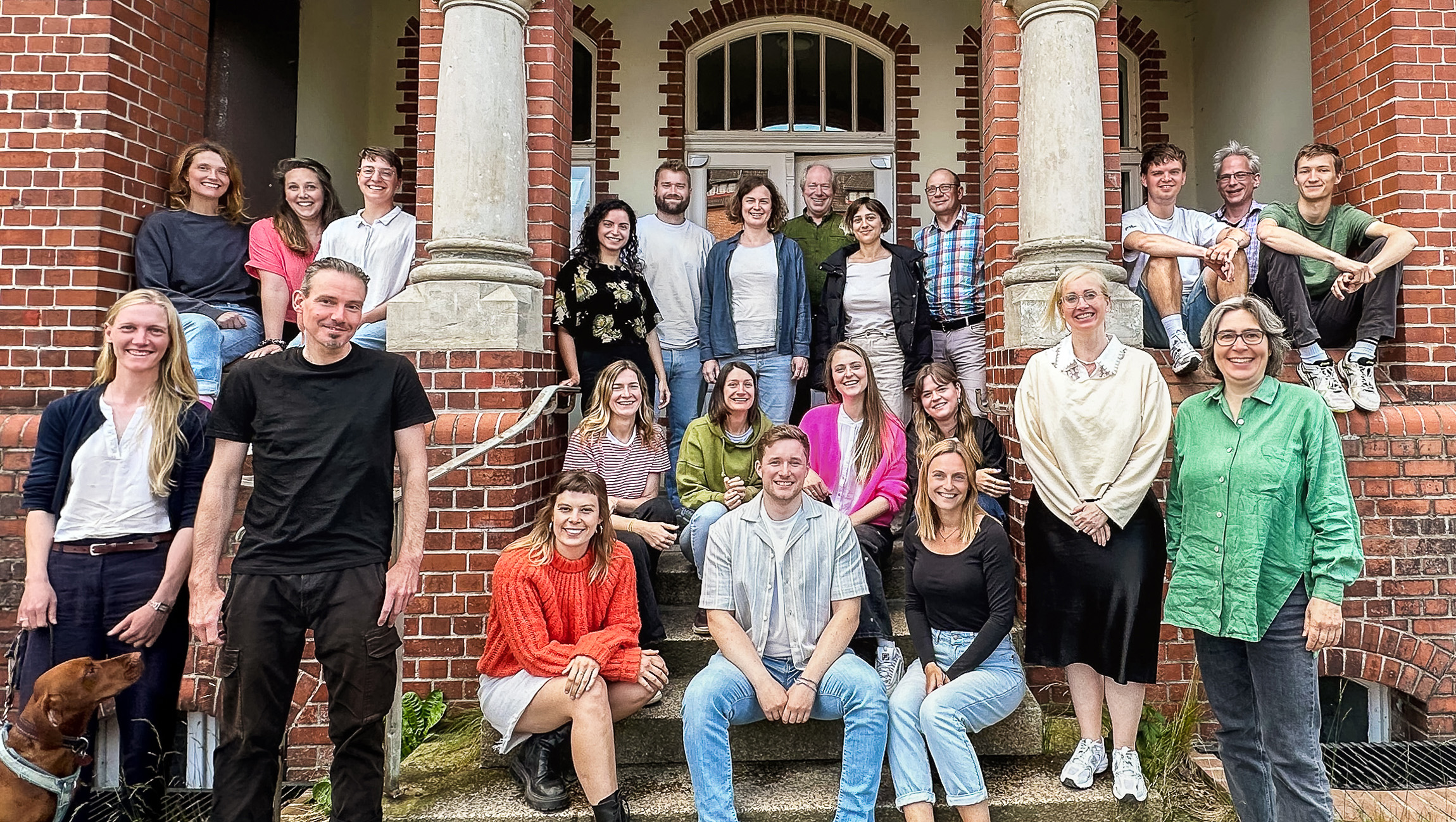 Teamfoto der Böll-SH. Etwa 20 Personen sitzen und stehen vor dem Eingang ihres historischen Bürogebäudes im Anschaspark Kiel.