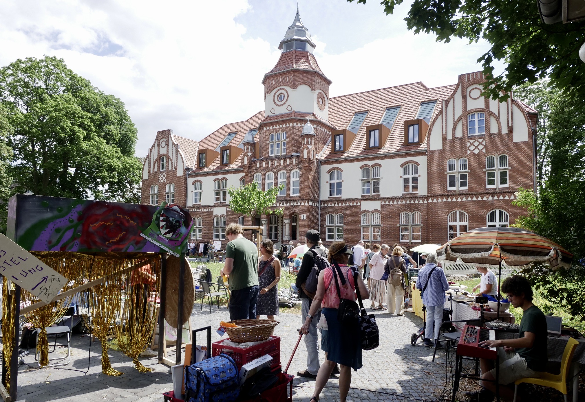 Ein historisches Gebäude im Anscharpark Kiel frontal fotografiert. Das Haus ist weiß und aus rotem Backstein. In der Mitte befindet sich ein Uhrenturm auf dem Dach des Hauses. Im Vordergrund ist ein Eisenzaun, im Hintergrund blauer Himmel mit Wolken zu sehen.