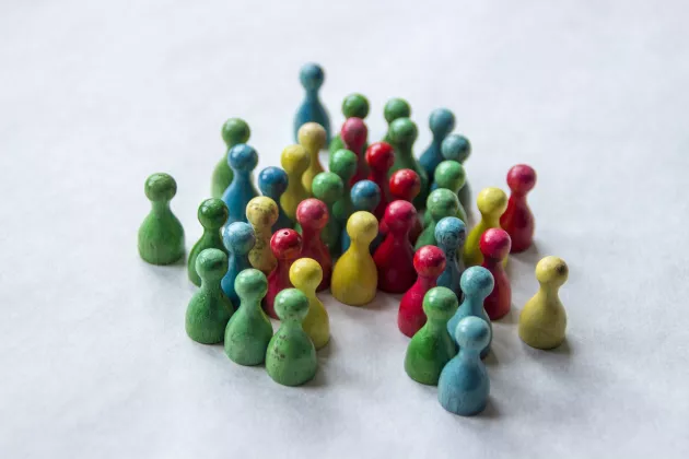 Kleine Spielfiguren aus Holz stehen in einer Gruppe zusammen. Sie sind blau, grün, rot und gelb.