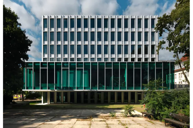 Zu sehen ist das Büro der Heinrich-Böll-Stiftung Berlin. Unten ist eine Glasfassade, darüber in mehreren Stockwerken eine weiße Fassade mit Fenstern. Links uns rechts stehen Bäume und Sträucher.