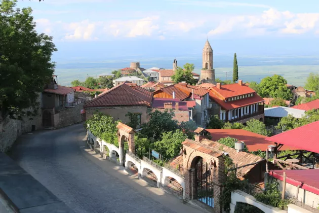 Ein Foto eines georgischen Dorfs. Vorne windet sich eine Straße, der Blick geht über rote Dächer im Grünen hin zu einem Horizont mit Wolken und blauem Himmel.