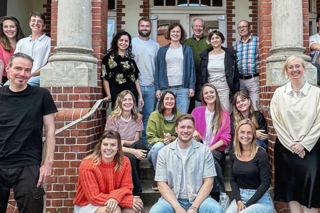Teamfoto der Böll-SH. Etwa 20 Personen sitzen und stehen vor dem Eingang ihres historischen Bürogebäudes im Anschaspark Kiel.