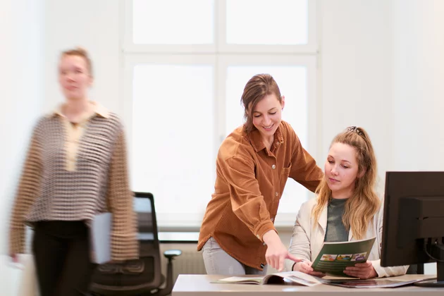 Eine Person sitzt am Schreibtisch und zeigt einer hinter ihr stehenden Person ein Heft. Im Hintergrund läuft eine weitere Person.