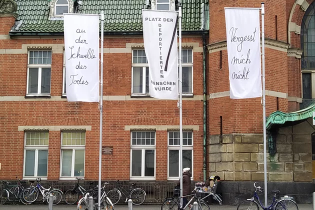 Vor dem Lübecker Hauptbahnhof hängen drei Flaggen anlässlich der gemeinsamen Landesgedenkstättentagung von Schleswig-Holstein und Mecklenburg-Vorpommern. Auf den Flaggen ist eine künstlerische Arbeit von Ute Friederike Jürßl abgedruckt.