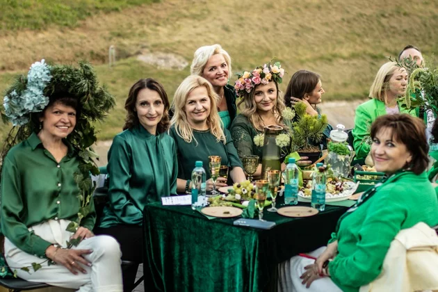 Mehrere Frauen sitzen beim Green Dinner im litauischen Alytus um einen gedeckten Tisch. Sie alle tragen grüne Kleidung und teilweise extravaganten Kopfschmuck aus Blumen und Gräsern.