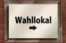 Im Hintergrund ist ein rotes Mauerwerk. Auf der Mauer ist ein Glasschild angebracht. In dem Schild hängt ein Papier auf dem steht: Wahllokal mit einem Pfeil, der nach rechts zeigt.