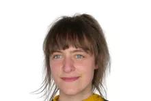  Porträt von Rieke Goetz. Eine junge Frau mit braunen Haaren in einem Pferdeschwanz gebunden und mit Pony lächelt in die Kamera. Sie trägt einen schwarzen Pullover und darunter einen gelben Rollkragenpullover.