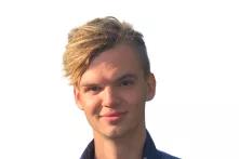 Porträt von Max Nettlau. Ein junger Mann mit kurzen blonden Haaren lächelt in die Kamera. Er trägt ein weißes Shirt mit einer blauen Jacke darüber.
