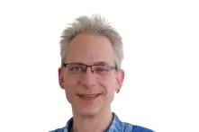 Porträt von Martin Petersen. Ein mittelalter Mann mit kurzen grauen Haaren lächelt in die Kamera. Er trägt eine Brille und ein blau-kariertes Hemd.