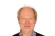 Porträt von Dirk Scheelje. Er hat kurze blonde Haare und lächelt in die Kamera. Er trägt einen dunklen Pullover mit einem hellen Hemd darunter.