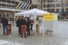 Auf einem Platz steht vorne eine Wahlkabine, auf der steht: Wähle hier symbolisch. Dahinter ist ein Pavillon aufgebaut, vor dem eine Gruppe von Menschen steht.