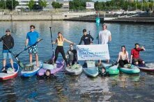 Auf dem Wasser liegen mehrere Stans-Up-Paddel-Boards nebeneinander. Auf den Boards stehen Leute mit Paddeln in der Hand. Zwei von ihnen halten ein Banner auf dem steht: Ocean Summit - jedena Tag für die Meere.