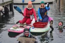 Zwei Personen sitzen in dicken Anzügen auf zwei SUPs und halten Gegenstände in den Händen. Im Wasser sind zwei Personen in Tauchanzügen.