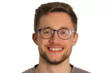 Porträt von Fynn-Ole Eisenhuth. Ein junger Mann mit kurzen dunkelblonden Haaren lächelt in die Kamera. Er trägt eine Brille, einen Dreitagebart und einen grauen Pullover.