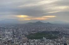 Panorama von Santiago de Chile. Viele Häuser stehen am Boden. Am Horizont sind Berge und wolkenverhangener grau-orangener Himmel.