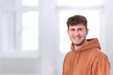 Portrait von Konstantin Gabler. Ein junger Mann mit kurzen braunen Haaren und einem Dreitagebart lächelt in die Kamera. Er trägt einen rostfarbenen Hoodie.