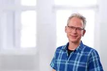 Portrait von Martin Petersen. Ein mittelalter Mann mit kurzen grauen Haaren lächelt in die Kamera. Er trägt eine Brille und ein blau-kariertes Hemd.