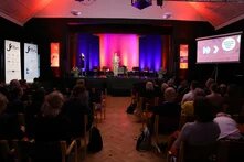 Im Hintergund steht eine Person auf einer in Rot und Blau beleuchteten Bühne. Vor der Bühne sitzen zahlreiche Personen auf Stühlen. Man sieht diese von hinten und blickt auf die Bühne.