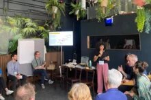 Eine Gruppe von Menschen sitzt zusammen auf Stühlen in einem Kreis. Sie hört einer Person zu, die frontal zu ihnen in ein Mikrofon spricht. Im Hintergrund befinden sich Poster- und Whiteboards sowie zahlreiche Pflanzen.