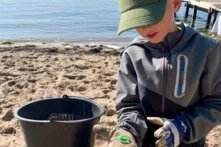 Ein Kind sitzt am Strand im Sand und schaut in seine Hand. Diese ist in einem Handschuh. Auf der Hand ist Plastikmüll, vermutlich ein Flaschendeckel, in Grün zu sehen. Neben dem Kind steht ein Eimer.