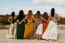 Sechs Frauen stehen nebeneinander im Arm mit dem Rücken zur Kamera zusammen. Sie halten ihre Hände hinter dem Rücken der jeweils neben ihnen stehenden Frau. Alle tragen Kleider. Im Hintergrund sind Dünen zu erkennen.