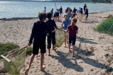 Ein Gruppe junger Kinder geht mit Keschern an einem Strand Richtung Wasser entlang. 