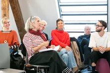 Eine Gruppe an Menschen sitzt auf Stühlen zusammen. Eine Person spricht. Die anderen hören ihr zu.