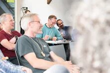 Vier Menschen sitzen auf Stühlen und scheinen einem Vortrag zu zu hören. Sie lächeln und schauen in eine Richtung.