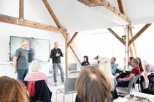 Zweo vortragende Personen stehen vor einem Bildschrim. Vor Ihnen sitzt eine Gruppe an Menschen.