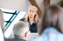 Es handelt sich hier um Jaako Bloomberg aus Finnland. Eine Person steht vor einer Gruppe Menschen und trägt etwas vor.