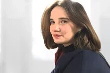 Portrait von Runa Schorge. Eine junge Frau mit brünetten, knapp über die Schulter langen Haaren. Sie trägt ein dunkelblaues Oberteil und sitzt seitlich zur Kamera.