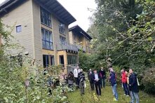 Eine Gruppe von Menschen steht vor einem Gebäude. Um sie herum sind Bäume und Büsche. Sie stehen auf dem Rasen.