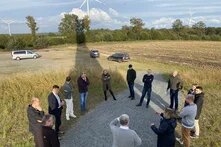 Eine Gruppe von Menschen steht im Halbkreis auf einem Kiesweg in einer ländlichen Umgebung. Im Hintergrund sind Windkraftanlagen und ein abgeerntetes Feld zu sehen. Einige Autos sind auf einem unbefestigten Parkplatz abgestellt. Die Personen scheinen sich im Gespräch zu befinden, während sie sich die Umgebung ansehen.