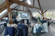 Eine Gruppe von Menschen sitzt in einem Vortrag in einem Halbkreis vor einem Bildschirm. Neben dem Bildschirm steht eine vortragende Person.