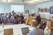 Vor einer Gruppe an Tischen sitzenden Menschen steht eine Person neben einer mit einem Beamer an die Wand projizierten Präsentation.