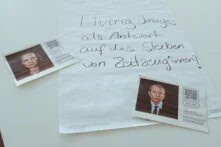 Zwei Zettel mit Personen darauf. Sie liegen zusammen mit einem großen Blatt Papier auf einem Tisch. Auf dem Blatt steht die Frage "Living Images als Antwort auf das Sterben von Zeitzeug*innen?"