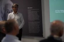 Eine Person steht vor einer Wand, auf der mit einem Beamer eine Präsentation projiziert wird. Vor der Person sitzen weitere Menschen.