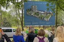 Personen stehen vor einem Bauplakat an der Alten MU in Kiel. Das Plakat zeigt, wie das Gebäude zu einem neuen, kreativen Dorf umgebaut werden soll.