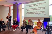 Einew Person hält einen Vortrag vor einer Leinwand, auf die eine Präsentation gebeamt wird. Es sitzen weitere Personen auf Stühlen neben der vortragenden Person.