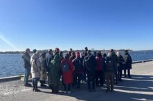 Eine Gruppe an Menschen steht vor einer Hafen-/Wasserkante und schaut Richtung Wasser.