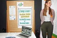 Eine Person steht neben einer Pinnwand. An der Pinnwand hängt ein Plakat mit der Aufschrift " Change School! Berufliche Schulen starten durch: nachhaltig lernen und Berufung finden"