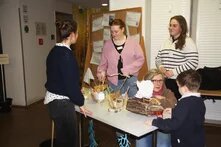 Vier erwachsene Personen befinden sich an einem Tisch. Ein kleines Kind steht vor einer vier erwachsenen Personen. Sie ist in der Hocke und spricht mit dem Kind. Auf dem Tisch stehen selbstgebaute Boote.