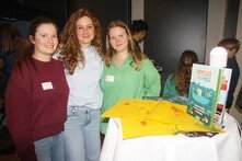 Drei Personen stehen zusammen Arm in Arm hinter einem Stehtisch. Sie scheinen ein Buch zu präsentieren, das auf dem Stehtisch steht.