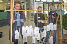 Drei jüngere Personen stehen hinter einem Gestell. An diesem sind verschiedene Kärtchen angehängt. Die Personen lesen/ schauen sich die Karten durch/ an.