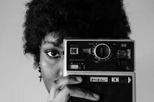 Schwarzweiß-Foto einer Schwarzen Frau, die eine alte Kamera vor ihr linkes Auge hält