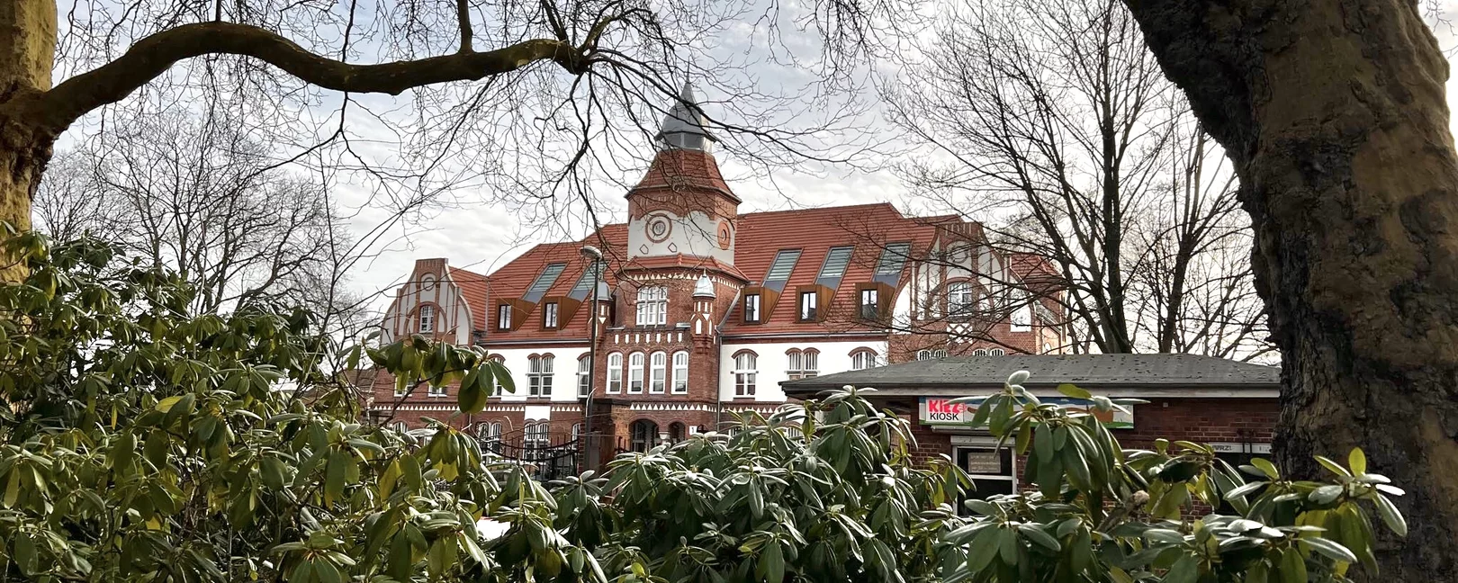 Haus 1 im Anscharpark Kiel. Ein historisches Backsteingebäude ist im Hintergrund. Davor sieht man hohe, grüne Sträucher und die Stämme von Bäumen.