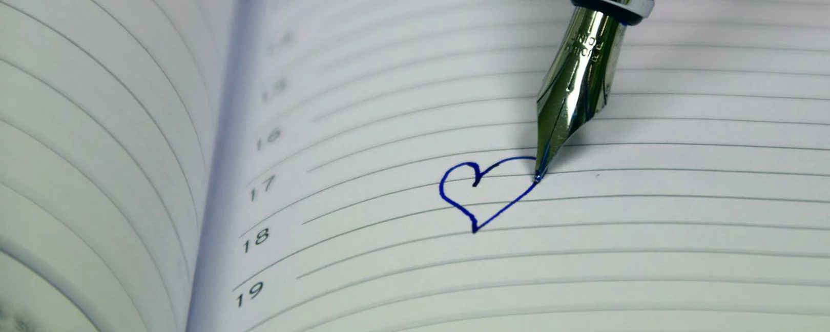 Man sieht Daten in einem Kalender. Die Spitze eines Füllers hat ein Herz auf das Kalenderblatt gemalt.
